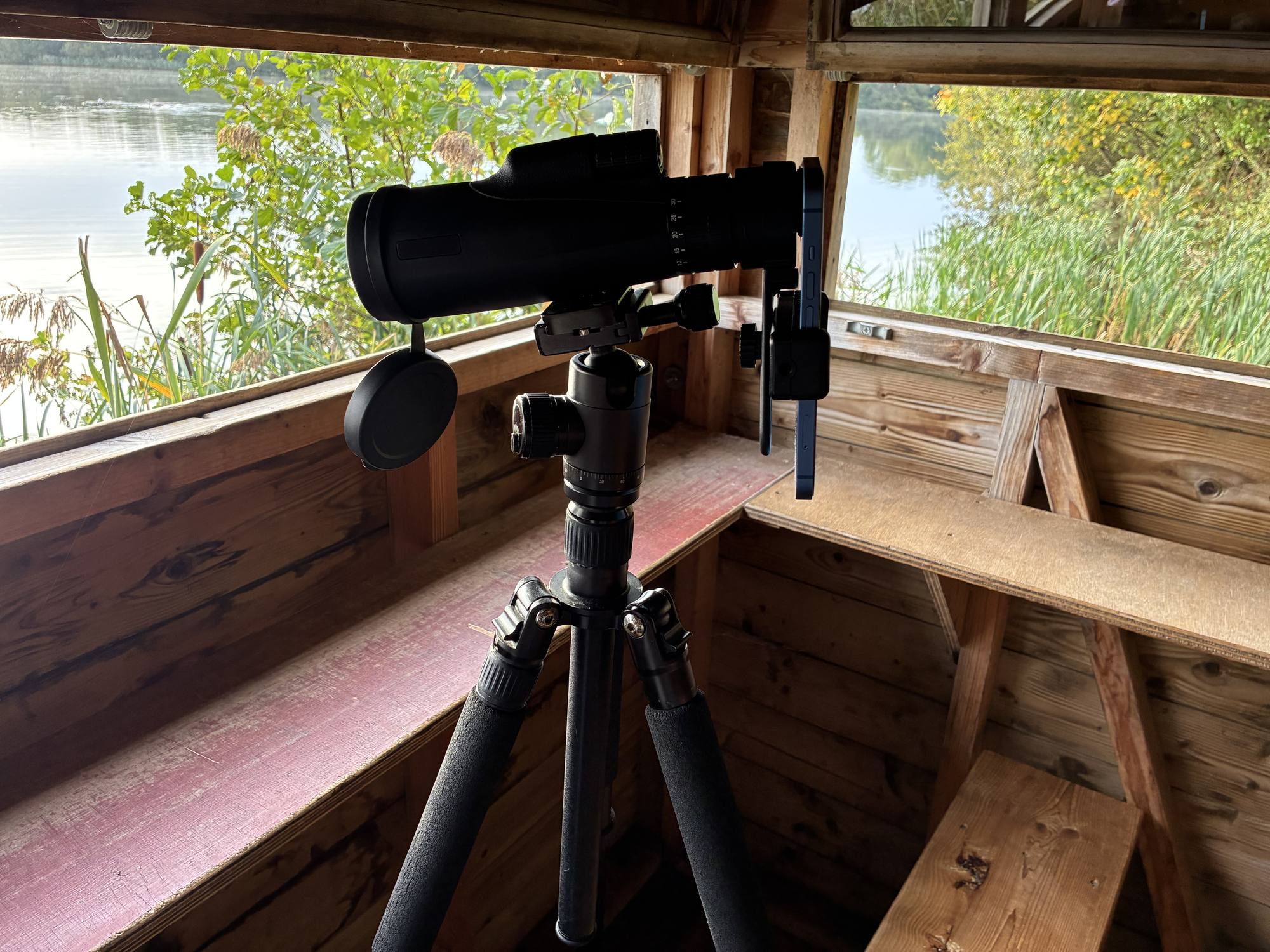 A phone held at a scope’s eyepiece in a bird hide, with a lake beyond.
